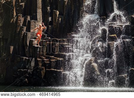 Middle-aged Man Jumping In Waterfall Lake. Falling Water Streams Flow On Black Volcanic Stone Cascad