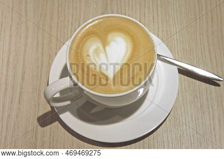 White Cup Of Coffee With Milk Foam On Wooden Table In A Cafe. Cup Of Hot Latte And Saucer With Spoon