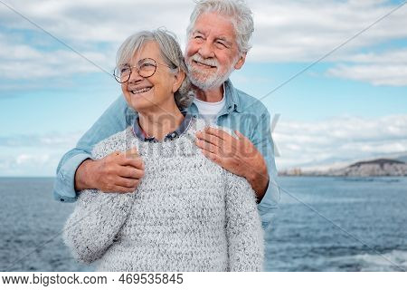 Happy Handsome Senior Couple Standing Embraced By The Sea Looking Away. Smiling Elderly Man And Woma