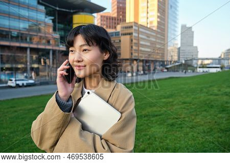 Stylish Smiling Girl, University Student In Trench Coat, Holds Tablet, Talks On Mobile Phone, Has Co