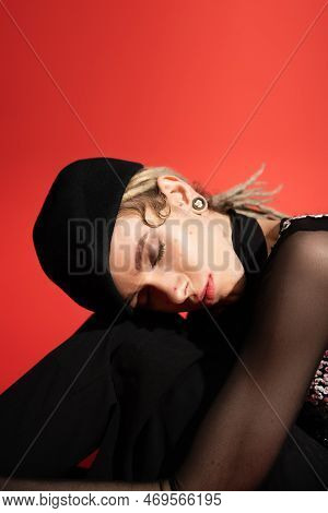 Portrait Of Queer Model In Black Beret Sitting With Closed Eyes Isolated On Red.