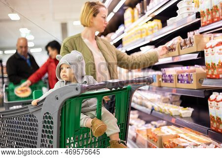 Casualy Dressed Mother Choosing Products In Department Of Supermarket Grocery Store With Her Infant 