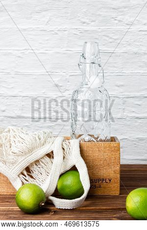 Lemonade Bottle In Crate With Green Limes On Wooden Table. Side View Of Fresh Citrus.