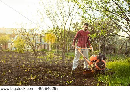 A Farmer Man Works In The Field, Plowing The Land With A Plow On The Farm. A Plowman On A Walk Behin