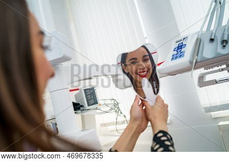 View From Side Of Happy Female Patient. Woman Looking At Mirror And Enjoying Beautiful Smile In Dent