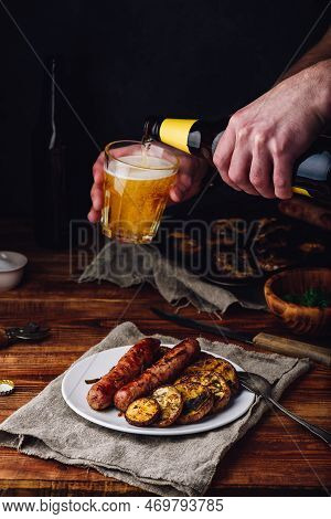 Pork Sausages Baked With Eggplant, Leek And Herbs On White Plate. Man Pouring Beer From Bottle Into 