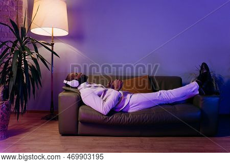 A Black Man Is Lying On The Sofa And Listening To Music. A Man In A White Suit Is Lying On A Green S