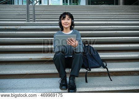 Young Woman Draws On Her Tablet, Listens To Music In Headphones. Asian Girl Sits On Stairs And Does 