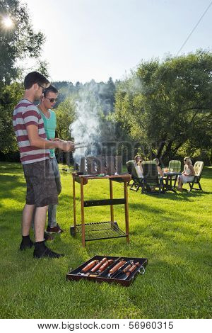 Group of friends enjoying a nice, warm, summer evening with food and drinks. Two men tending to a barbecue, with a group of their friends sitting around a picnic table in the background