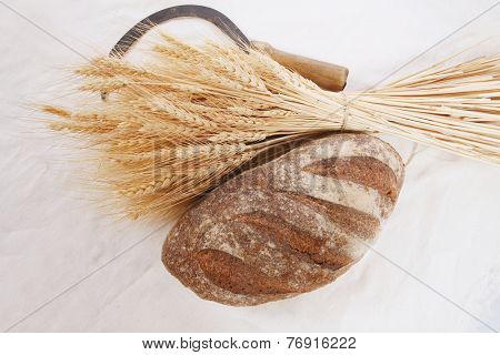 bunch of mown wheat ears with vintage handmade reaper hook sickle and rye dark french fresh bread loaf on white linen tablecloth