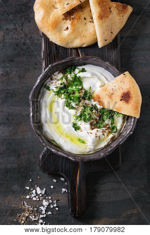 labneh middle eastern lebanese cream cheese dip with olive oil, salt, herbs served traditional pita bread in terracotta bowl over dark texture metal background. Top view with space