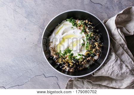 labneh middle eastern lebanese cream cheese dip with olive oil, salt, herbs, olives tapenade served in black bowl with textile napkin over gray texture metal background. Top view with space