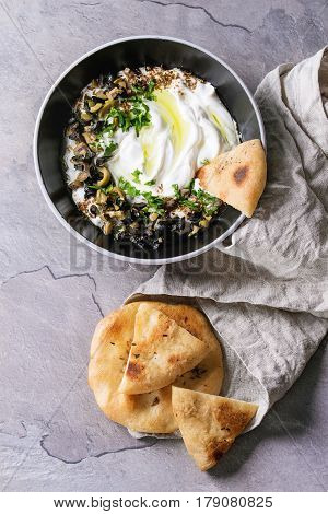 labneh middle eastern lebanese cream cheese dip with olive oil, salt, herbs, olives tapenade served in black bowl with traditional pita bread over gray texture metal background. Top view with space