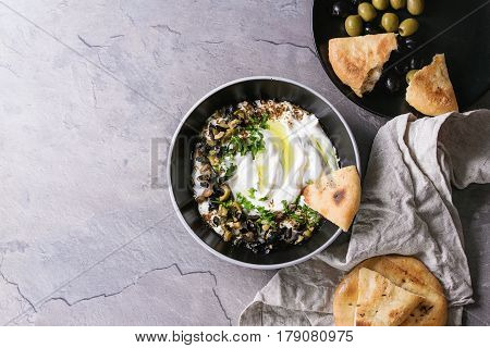 labneh middle eastern lebanese cream cheese dip with olive oil, salt, herbs, olives tapenade served in black bowl with traditional pita bread over gray texture metal background. Top view with space