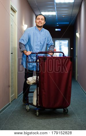 Young man pushing a housekeeping cart laden with clean towels, laundry and cleaning equipment in a hotel as he services the rooms.