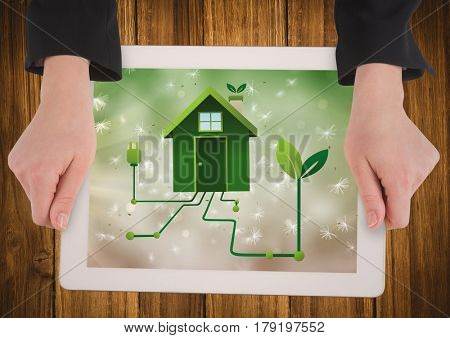 Digital composite of Hands with tablet on table showing green house graphic against bokeh