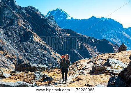 One Person with Backpacks and trekking Poles walking up on Mountain Footpath with mixed rocks and grass terrain with high Cliffs and Peaks beside