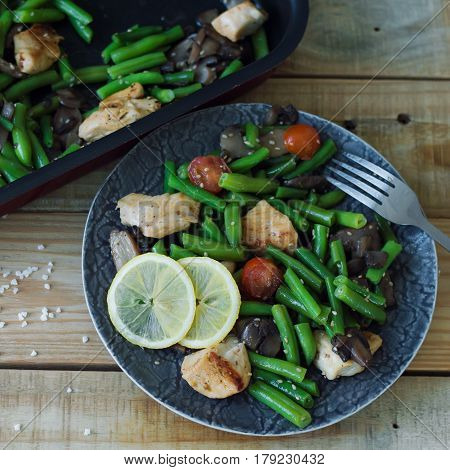 Square photo of hot salad with green beans baked cherry tomatoes chicken breast sliced mushrooms sesame seeds and lemon served in a grey plate with metal fork on wooden board