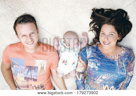 Family portrait of a happy young couple lying down on the floor with their newborn baby sleeping in the middle.