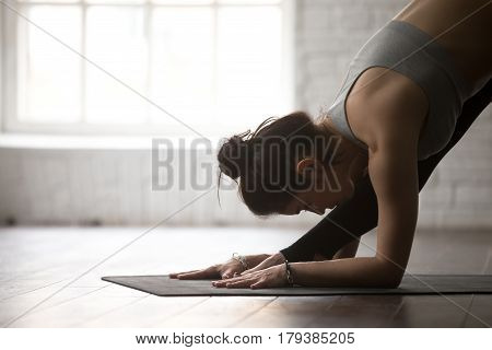 Young attractive yogi woman practicing yoga concept, stretching in One Sided Fold, Ardha Parsvottanasana exercise, Half Pyramid pose, working out, wearing black sportswear, white loft studio, close up