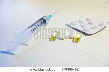 A variety of medication including a syringe with a green tip blister pack white pills and yellow plastic pills full of cod liver oil vitamin D supplement shot at a low angle against a white background