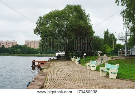 Small cozy promenade with tratuar and wooden pier for boats on the lake or river in a green park in the city. Quiet summer cloudy day in the park by the lake.