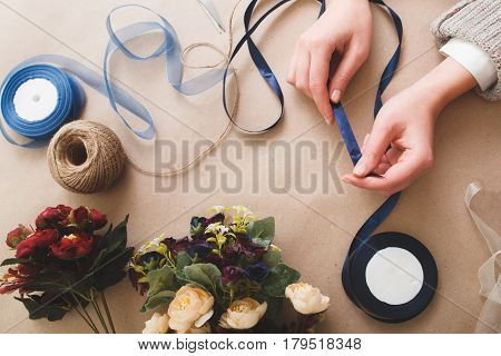Preparing decorations for holiday, fancywork, handmade art. Unrecognizable woman hold blue ribbon on beige background with artificial flowers, tape rolls and rope, flat lay.