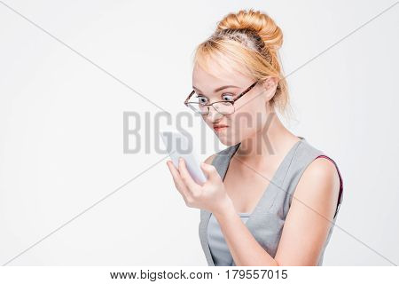 Young woman in glasses with mobile phone looks angry, peeved and irritated. Annoyed and grumpy blonde with smartphone on grey background with copy space.