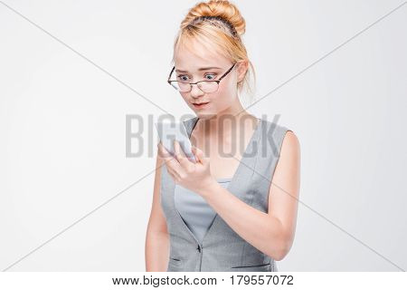 Young woman in glasses with mobile phone looks angry, peeved and irritated. Annoyed and grumpy blonde with smartphone on grey background with copy space.