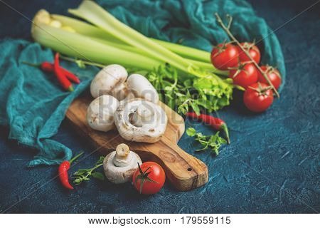 Fresh mushrooms champignons on a wooden cutting board parsley greens celery stalks cherry tomatoes and hot red chili pepper on a dark blue background