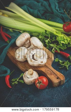 Fresh mushrooms champignons on a wooden cutting board parsley greens celery stalks cherry tomatoes and hot red chili pepper on a dark blue background