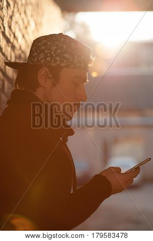 young man in a baseball cap holds a smartphone and looks at it against a brick wall background lit by the backlight with lens flare