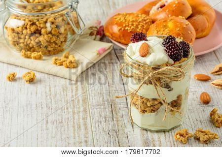 Healthy breakfast ingredients. Homemade granola with yoghurt in open glass jar and fresh fragrant buns. The food is rich in useful carbohydrates vitamins and nutrients that are essential for health. The dishes stand on a white wooden table. Copy space.
