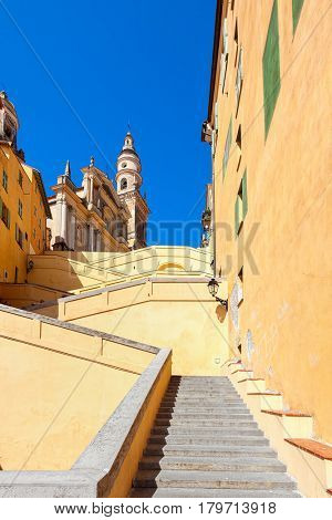 Stairs among colorful walls towards Saint-Michel Archange Basilica under blue sky in Menton, France (vertical composition).