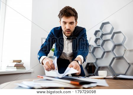 Absentminded young businessman in suit rummaging in papers at workplace, office background.
