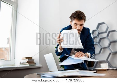 Absentminded young businessman in suit rummaging in papers at workplace, office background.