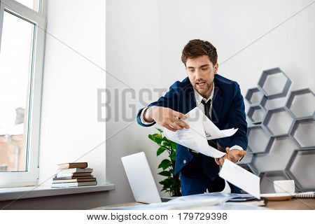 Absentminded young businessman in suit rummaging in papers at workplace, office background.