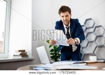 Annoyed young businessman in suit rummaging in papers at workplace, office background.