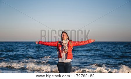 Young happy girl with backpack standing on a seaside breathing fresh air raising arms with eyes closed