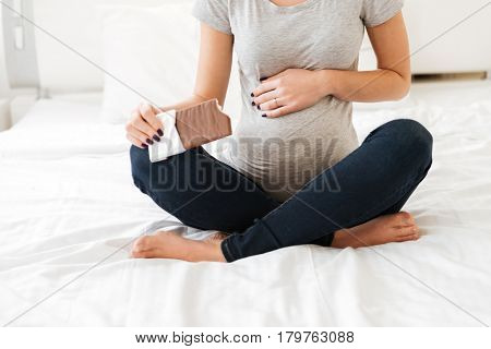 Closeup of pregnant young woman sitting with legs crossed and eating chocolate bar on bed in bedroom