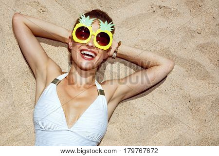 Smiling Woman In Swimsuit And Pineapple Glasses Laying On Sand