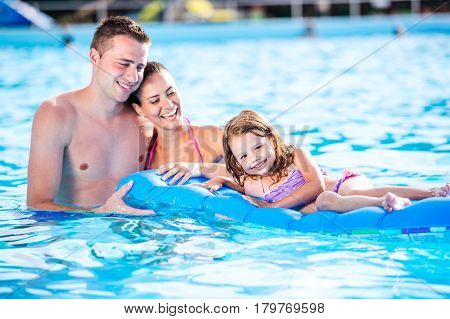 Young mother and father with their daughter sitting on pool lilo in swimming pool in aqua park. Summer heat and water.