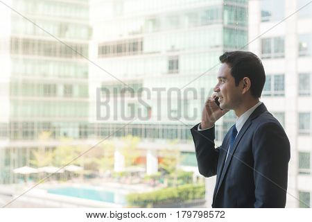 Mature and confident business executive looking looking out of large windows at a view of the city below from the top floor of an office building while talking on his mobile phone