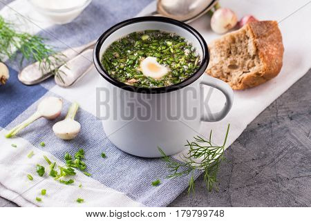 Traditional russian Cold Soup Okroshka with meat, vegetables and kvass serving size in enamel cup and raw ingredients on on grey stone table. Kvass is a traditional Slavic and Baltic fermented beverage commonly made from rye bread