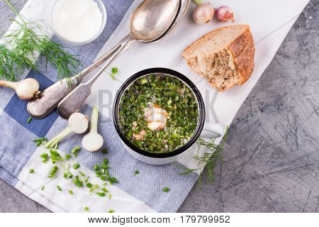 Traditional russian Cold Soup Okroshka with meat, vegetables and kvass serving size in enamel cup and raw ingredients on on grey stone table. Kvass is a traditional Slavic and Baltic fermented beverage commonly made from rye bread