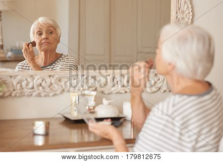 Senior woman applying foundation to her cheek with a makeup brush while sitting alone in front of a mirror in her bedroom at home