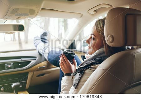 Woman traveler sitting in a car on passengers seat with feet in warm socks on car dashboard. Drinking take away coffee on road. Road trip. Freedom travel concept. Sun light flair effect