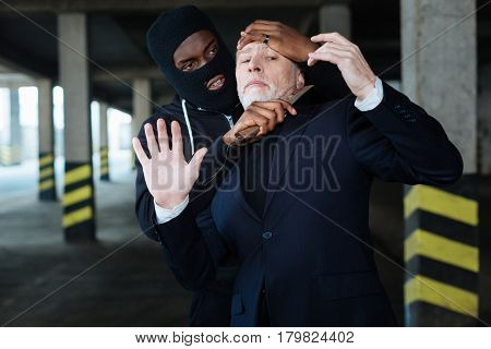 Aggressive behaviour. Aggressive ruthless male criminal wearing a mask and holding a knife while trying to kill a businessman
