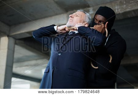 Being a murderer. Cruel ruthless serious criminal standing behind the businessman and killing him while using the rope