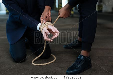 You will not escape. Dangerous afro american male criminal standing above his victim and binding his hands while holding him hostage